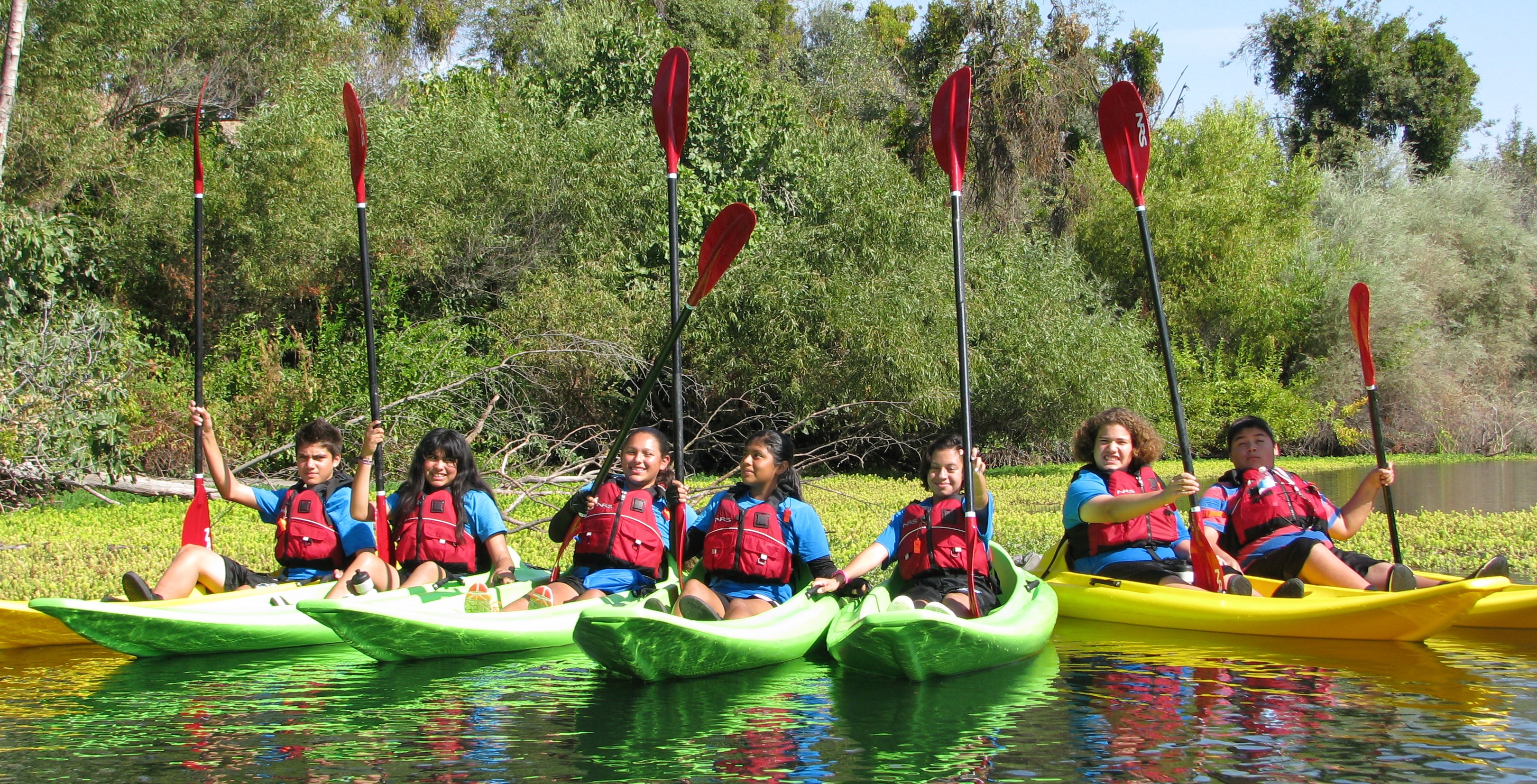 Photo of seven teenagers lined up next to each other each in a kayak on calm water near a tree and shrub lined shore smiling at the camera. Each holds their paddle upright so it stands in the air like a flagpole. 